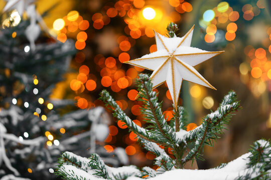 Christmas tree decorated with white star, ornaments and warm fairy lights, displayed outdoors with festive illuminated, light bokeh background and snowy branches. Holiday atmosphere, aesthetic mood
