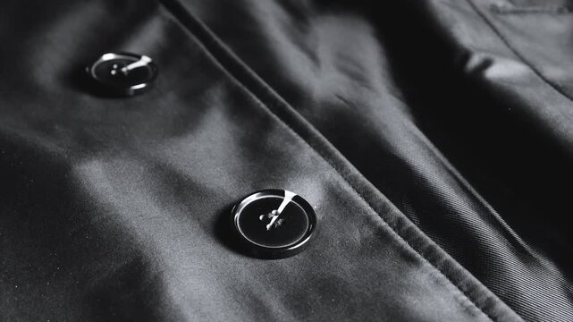 A close-up of a black satin trench coat with large round buttons showcases smooth waterproof fabric, neat seams, and elegant structural details of classic outerwear.
