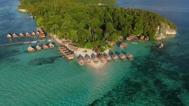 Aerial view of Raja Ampat overwater bungalows on stilts, white sand beach, lush tropical vegetation, and turquoise coral reef waters in Kri Island, Southwest Papua, Indonesia.