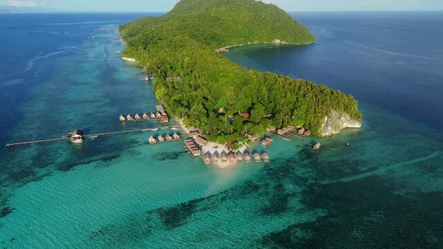 Aerial view of Raja Ampat Regency featuring overwater bungalows and lush tropical forests surrounded by turquoise coral reefs in Kri Island, Southwest Papua, Indonesia.