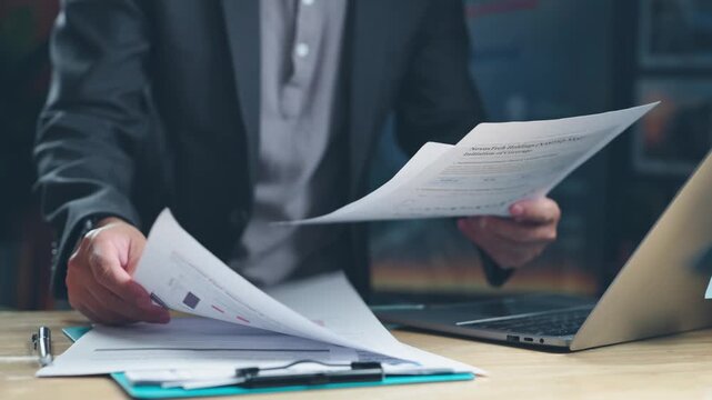 Focused businessman holding financial report documents above wooden desk with laptop computer, reviewing market research data, investment analytics, sales statistics, corporate balance sheet figures.