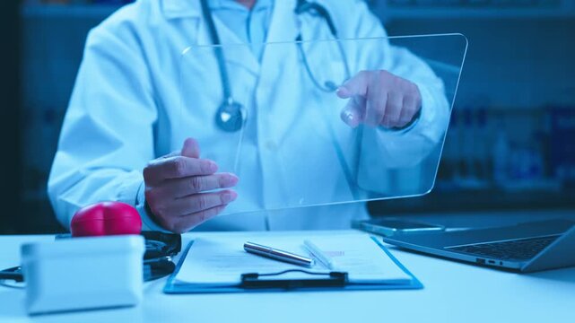 Focused doctor interacts with clear digital tablet screen seated behind clinic desk with stethoscope, clipboard paperwork, laptop, red toy heart, checking electronic medical chart records indoors.