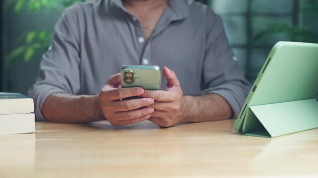 Midsection view casual man holding mobile phone at wooden desk near tablet computer stack books indoors, browsing social media text messaging in home office.