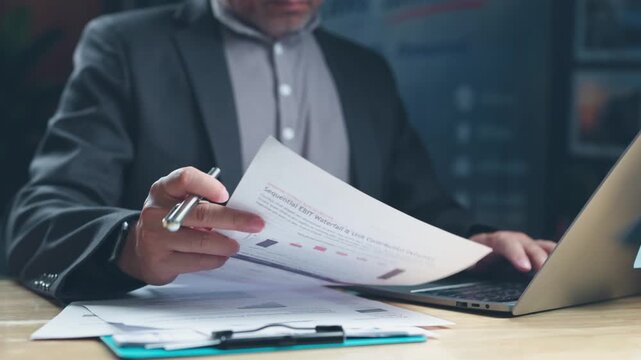 Focused mature businessman analyzing financial report sheets holds silver pen while typing on laptop keyboard at desk inside corporate office.
