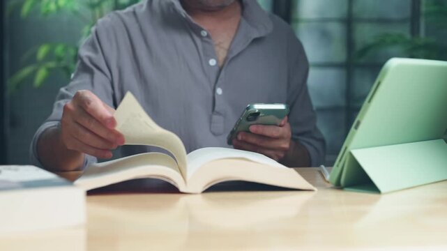 Focused man turning page open book on wooden desk beside digital tablet mobile phone in home study room, multitasking between printed literature modern communication tools.