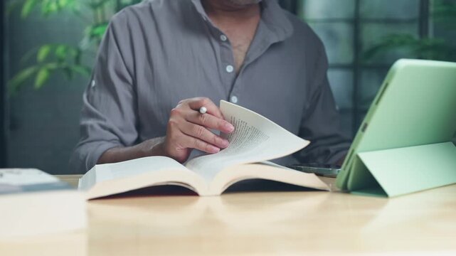 Focused man studying at wooden desk holding stylus while reading open printed book beside digital tablet screen indoor.