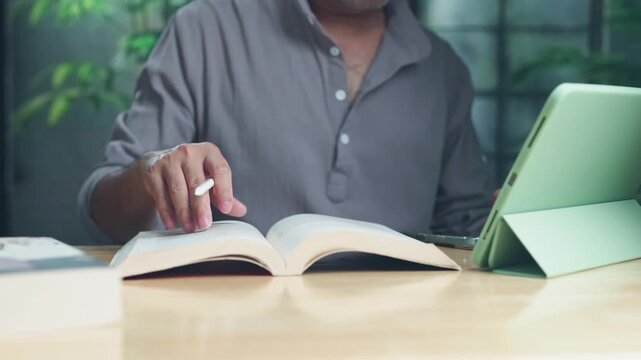 Focused man holding stylus over open book near digital tablet at wooden desk prepares research notes indoors during quiet afternoon study session.
