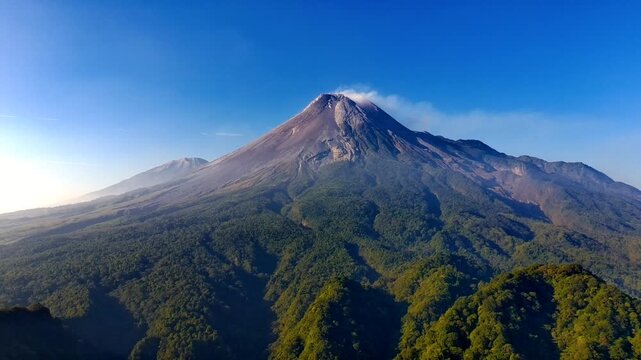 Aerial view of Gunung Merapi volcano with lush green forested slopes and a clear blue sky in Kecamatan Pakem, Daerah Istimewa Yogya, Indonesia.