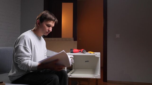 Man sitting on sofa intently reading flat pack furniture instructions while preparing tools and parts for diy assembly in a modern apartment living room during a move renovation project