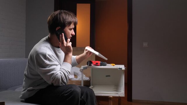 Young man holding a mobile phone to his ear, getting advice or support while struggling with flat pack furniture assembly using a drill and small parts at home