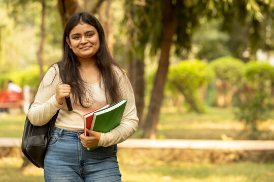 Portrait of indian young girl college student with backpack and notebooks standing and smiling in park