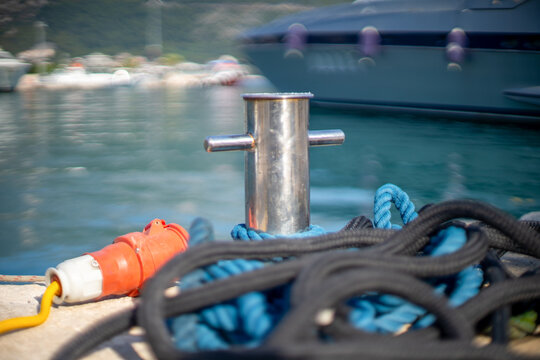 A stainless steel mooring bollard holds coiled blue and black rope on a marina dock, with an orange electrical connector nearby and a yacht docked in the harbor behind.