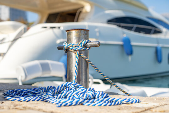 A blue and white striped rope is knotted around a stainless steel mooring bollard on a dock, with a luxury yacht moored in the background at a marina.
