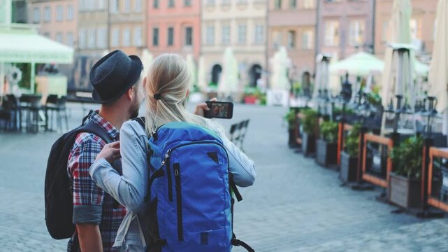 A man and woman tourists with backpacks taking photos on smartphones in a European city square with outdoor cafes and pastel buildings during daytime.