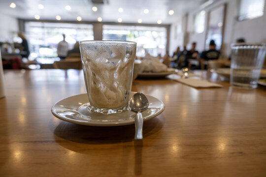 A finished coffee glass coated with leftover cream residue on a caf&eacute; table, with a blurred indoor dining interior and customers sitting in the background. An everyday, unposed moment after breakfast
