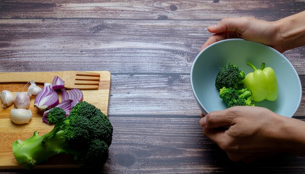 木製テーブルで新鮮な野菜を調味する様子（ブロッコリー, 赤玉ねぎ, パプリカ, ニンニク, まな板, 健康食, 料理教室, 背景素材） / Fresh vegetables and hands holding bowl on wooden table