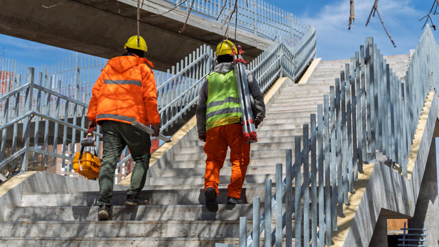 Two surveyors carrying topographic equipment and tripod climbing concrete stairs on a pedestrian bridge

