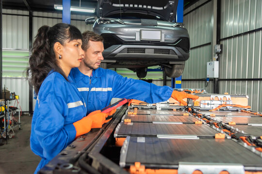 Electric vehicle technicians inspect high-voltage EV battery pack using a digital tablet in automotive workshop, showing battery diagnostics, maintenance, teamwork and engineering technology.