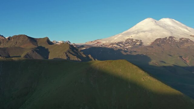 Aerial view of the snow-capped Mount Elbrus and the surrounding Caucasus Mountains with green slopes under a clear blue sky in Russia.