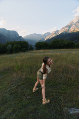 Obraz premium Young woman in a light hiking outfit stands in a wide grassy field with towering mountains in the background, enjoying a carefree outdoor moment.