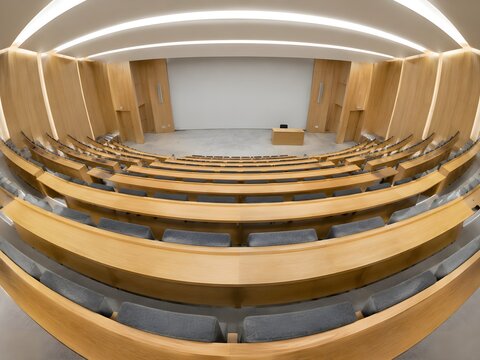 Raked tiers of empty lecture hall bench seating in honey oak leading down to a bare teaching wall