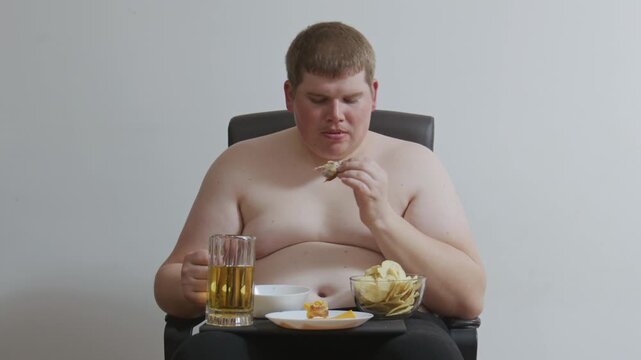 Overweight shirtless man eating junk food and drinking beer. Concept of obesity, sedentary lifestyle, bad food habits. Obese single male sitting in a chair and binge eating chips with alcohol.