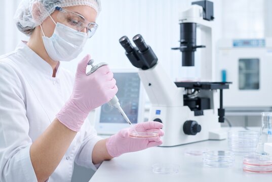 Professional female embryologist in protective gear using a micropipette and Petri dish for IVF research in a modern sterile laboratory