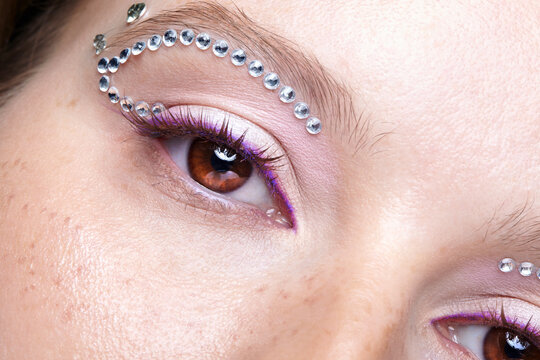 Extreme closeup of woman brown eye with rhinestone brow decorations and violet eyeshadow makeup.