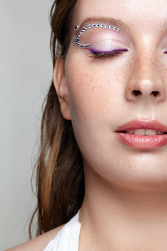 Close up portrait of woman face with closed eye, purple eyeliner, rhinestones and freckles on cheek.