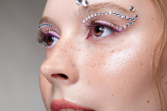 Closeup portrai of female with freckles on cheek, rhinestone eye makeup and purple eyeliner.