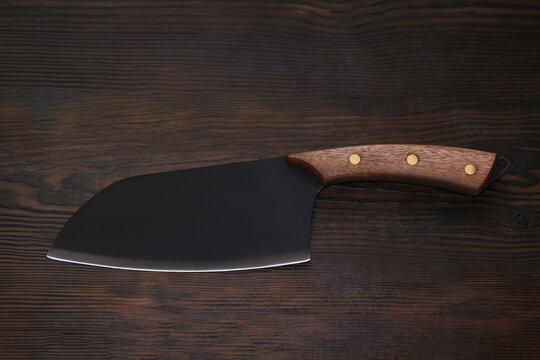 Professional chef meat cleaver with sharp black blade and wooden handle lying on dark textured wooden table background. Cooking concept.