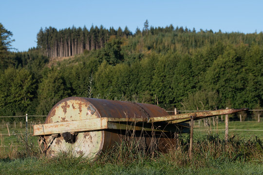 A large metal roller stands in a pasture surrounded by greenery. The old agricultural roller is rusty and overgrown with grass.