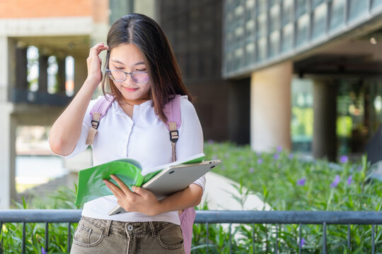Focused asian female student reading a book and holding tablet on campus, young university student wearing glasses studying with textbook and computer device outdoors