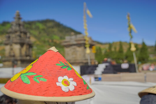 A close-up of a colorful hand-painted traditional conical bamboo hat with flower patterns, with ancient Hindu temples under a clear blue sky blur in the background during the Dieng Culture Festival.
