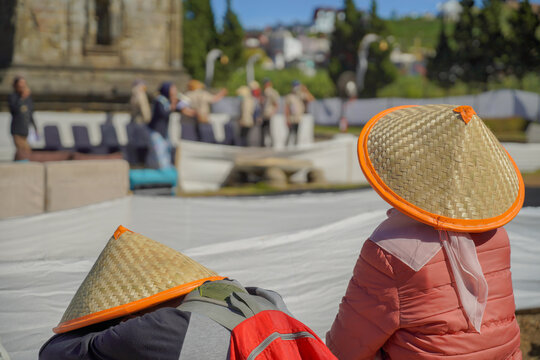 People wearing traditional conical bamboo hats with orange trim are sitting outdoors, watching an event or ceremony near an ancient stone temple structure during DIENG CULTURE FESTIVAL.