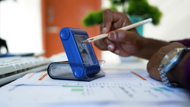 Person using a stylus on a handheld electronic device, Close-up of a person using a stylus on a blue handheld electronic device beside eyeglasses and printed documents on a desk.