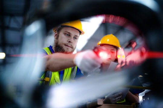 Two warehouse employees, a bearded man and a young woman, wearing yellow hard hats align a laser tool across the production tooling inside a car parts manufacturing facility with metal.