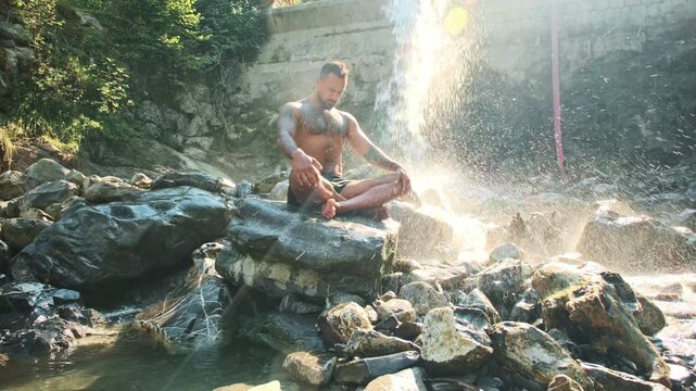 Yoga man breathing beside a waterfall. Man practice yoga with mindful breathing outdoors. Breathing exercises during yoga in nature. Breathing meditation in seated yoga poses.