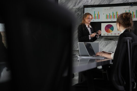With an open-handed gesture at the pie chart, the presenting businesswoman explains proportions to her colleagues seated in the shadows of the marble boardroom in a bright modern coworking.