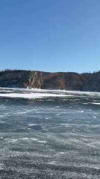 Slow motion vertical video of car driving on ice road of frozen Baikal Lake. View from cabin on ice surface and rocky coast of Olkhon Island on sunny winter cold day. Auto travel and adventure