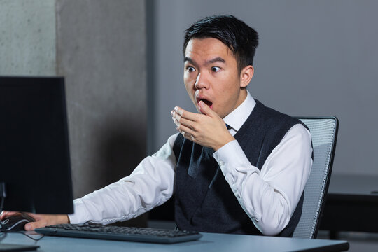 A surprised young Asian office worker covers his mouth while reacting to something on a computer, for workplace surprise, unexpected news, or emotional reaction concepts.
