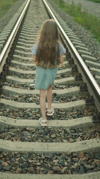 Long haired child in casual clothing steadily moves along parallel train tracks. scene is set outdoors with gravel and greenery visible around rails. Lighting suggests daytime creating calm