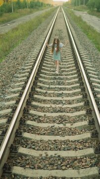 Young girl walks alone on railway track moving steadily along parallel rails. surrounding area features grassy fields or paths beside train tracks with scattered vegetation and gravel