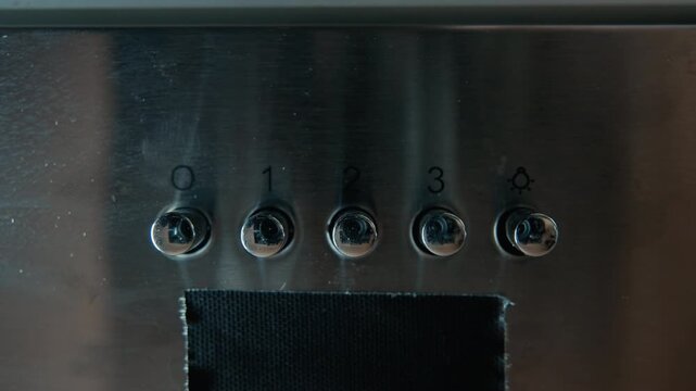 Straight-on close view of stainless steel control panel with five numbered metal push buttons aligned horizontally above black grille, while fingertip presses rightmost button and moves toward surface