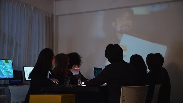 A group of professionals attends a video conference presentation in a dark office at night, watching a speaker on a large projection screen while collaborating on work together.