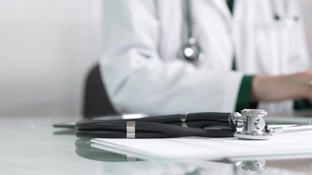 Stethoscope and medication history records form are lying on a glass table before a doctor working with laptop in clinical office background. Medicine concept