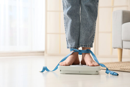 Weight loss and body image struggle, person standing on bathroom scale with measuring tape tied around ankles in home interior