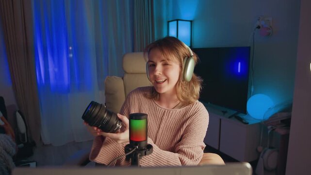 Young woman wearing over-ear headphones holds camera with both hands and speaks toward smartphone mounted on desk microphone, suggesting live tutorial, review, or creative presentation