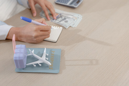 Travel savings concept. hands writing budget notes while counting cash beside passport, calculator, toy plane, and suitcase on a desk. travel, savings, budget, money, vacation.