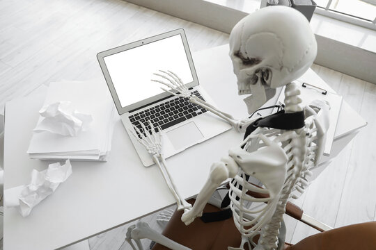 Burnout and workplace stress, human skeleton working on a laptop at an office desk with crumpled paper corporate fatigue and job pressure